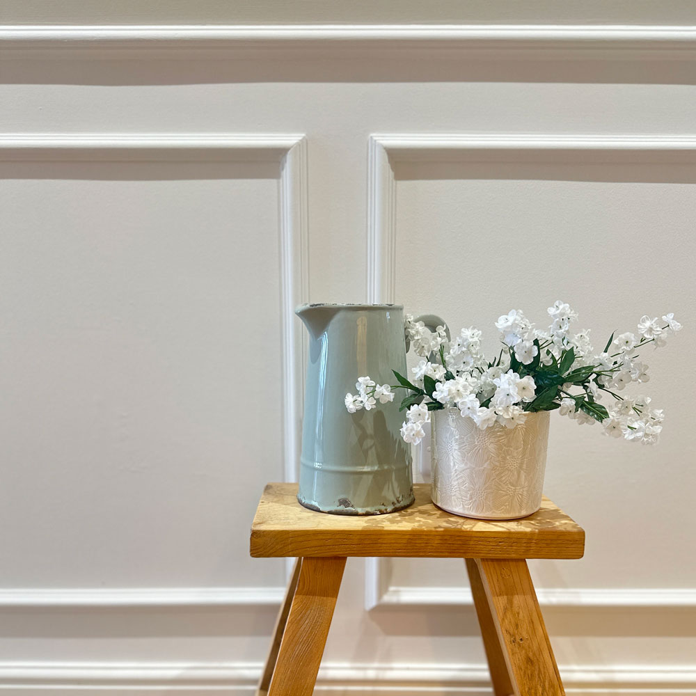 Traditional wainscot wall panelling detail in a neutral hallway with decorative moulding and a wooden stool styled with a jug and white flowers.